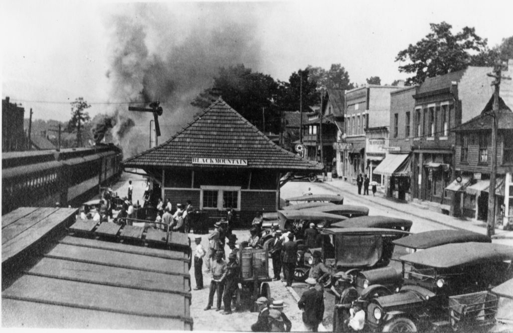 A train arrives at the Black Mountain railroad station on Sutton Avenue People and cars wait for the train. Store fronts line the road to the north of the station